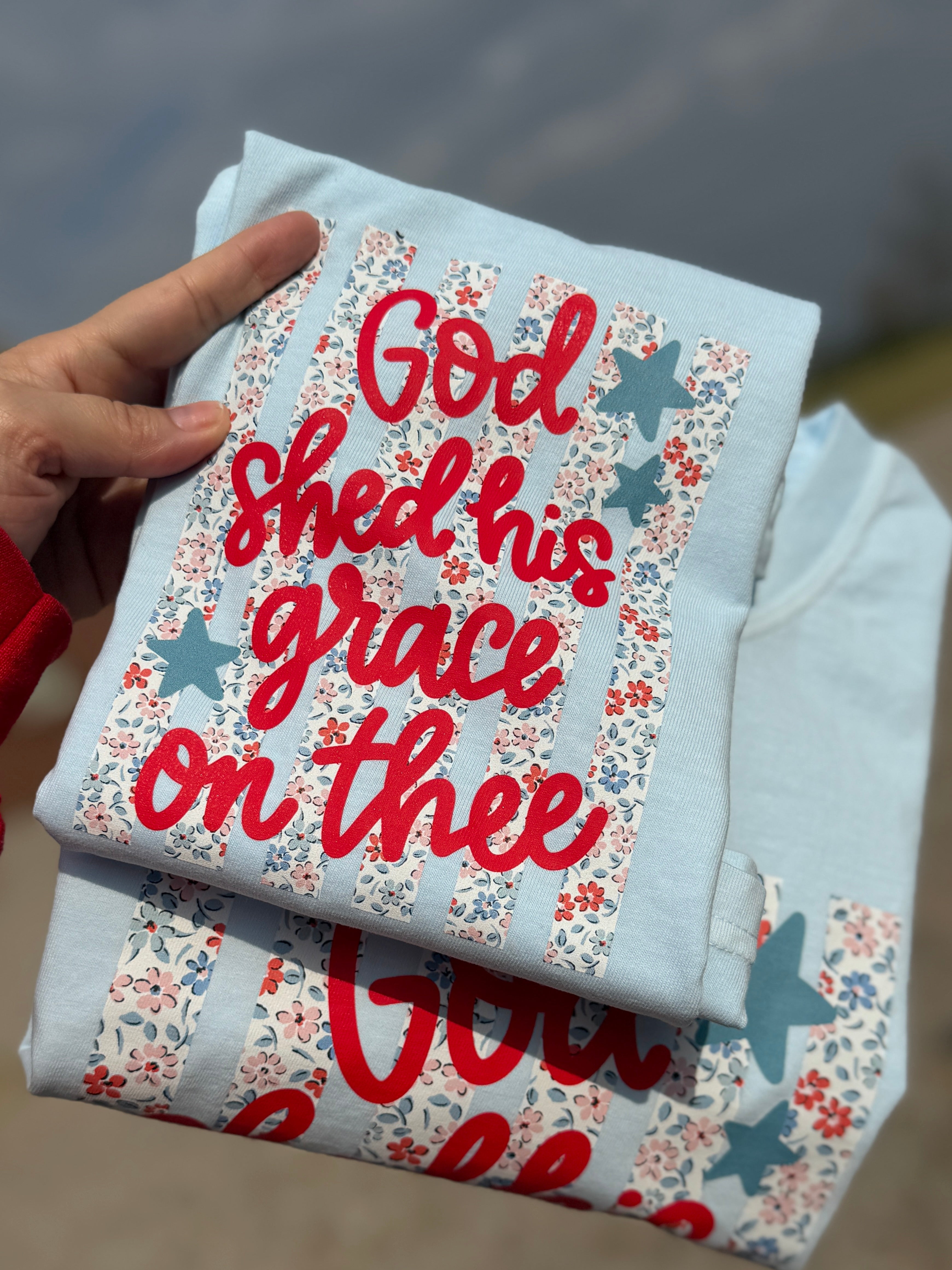 Light blue Christian graphic t-shirt with floral American flag design and red lettering that reads “God sheds His grace on thee”
