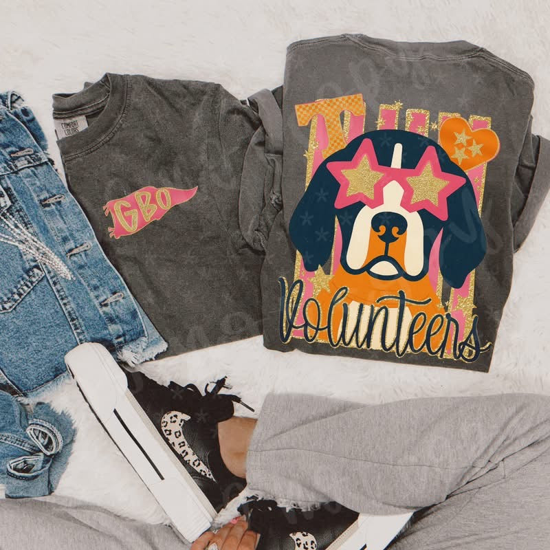 Flat lay of a trendy Tennessee Volunteers graphic t-shirt on a soft neutral background. The Comfort Colors pepper tee features a small left chest “Vols” logo and a bold back design with a stylish dog wearing star sunglasses in orange, pink, and gold tones. The shirt is styled with distressed denim jeans, black sneakers, and cozy loungewear pieces, creating a casual, game day outfit perfect for fall football season.